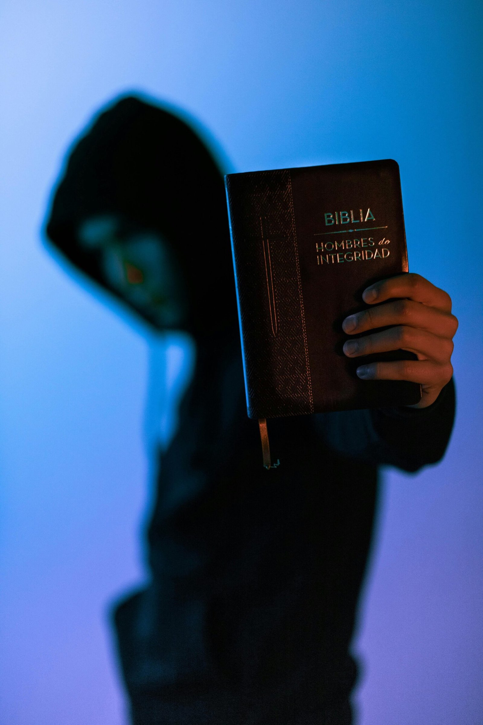 Hooded figure holding a Bible against a vibrant blue background, capturing a sense of mystery.